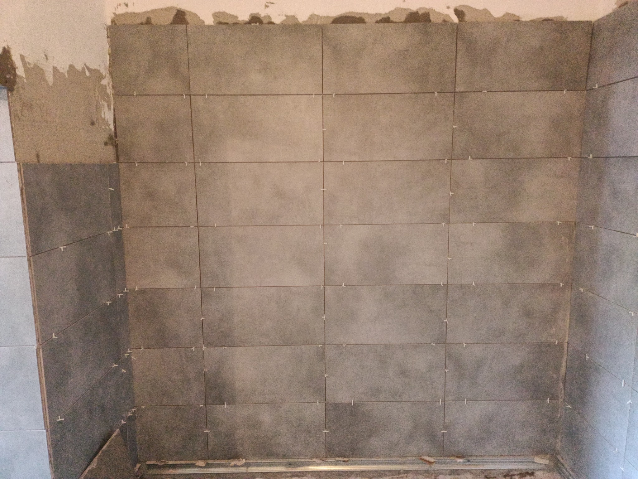 Ściana w trakcie układania szarych płytek ceramicznych, widoczne krzyżyki dystansowe i fragment surowej ściany z resztkami zaprawy.