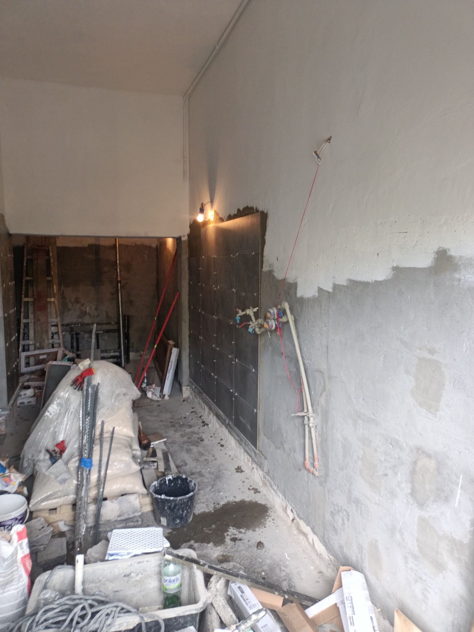 Wnętrze w trakcie remontu: częściowo ułożone ciemne płytki na ścianie, widoczne rury instalacji wodnej, surowe ściany z widocznymi śladami po gładzi, na podłodze bałagan, worki z materiałami...