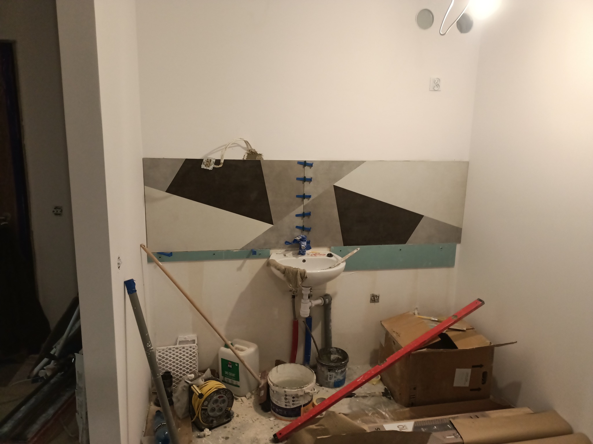 Łazienka w trakcie remontu: ułożone płytki z geometrycznym wzorem nad umywalką, niebieskie krzyżyki dystansowe, widoczne narzędzia i materiały budowlane, bałagan na podłodze.