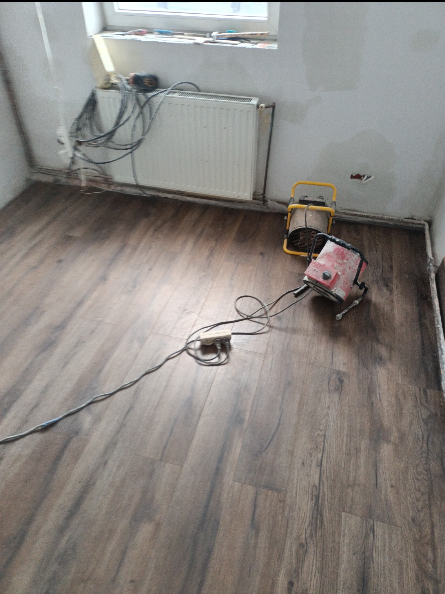 Pokój w trakcie remontu: nowa podłoga laminowana, grzejnik, ściany przygotowane do malowania, dwa grzejniki elektryczne. Kabel zasilający leży na podłodze.