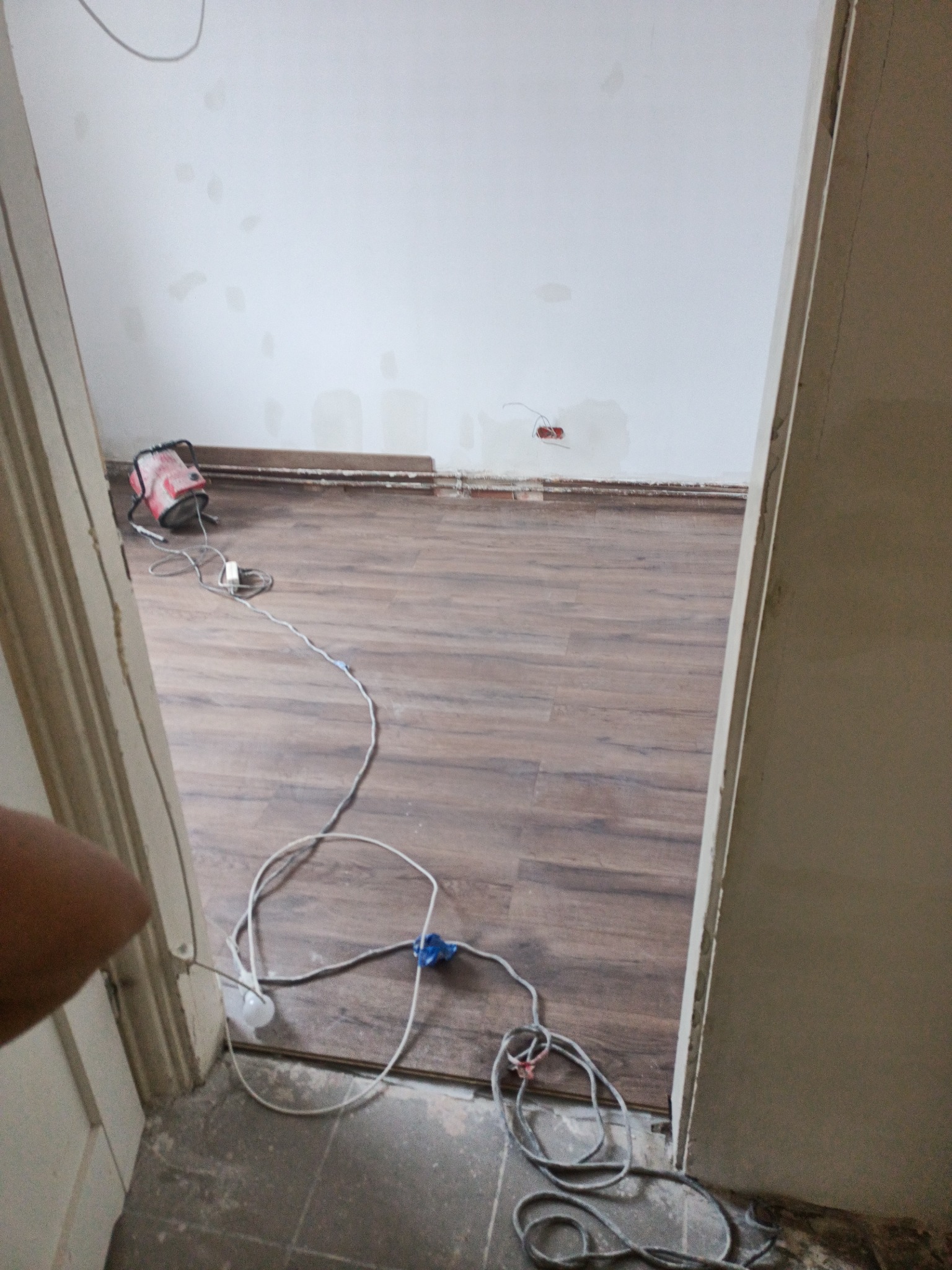 Przygotowanie do prac wykończeniowych: widok na podłogę z panelami, ściany z widocznymi śladami po szpachlowaniu, przewody elektryczne i lampa halogenowa na podłodze w trakcie prac.