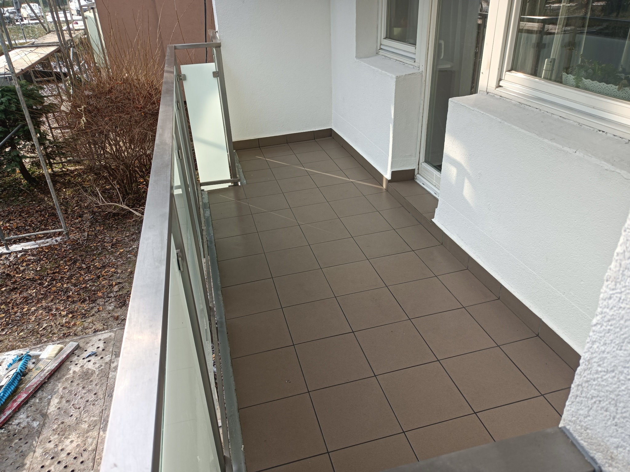 Wykafelkowany balkon z metalową balustradą i przeszklonym fragmentem, widok z góry na brązowe płytki, białe ściany i fragment rusztowania z narzędziami po lewej stronie.