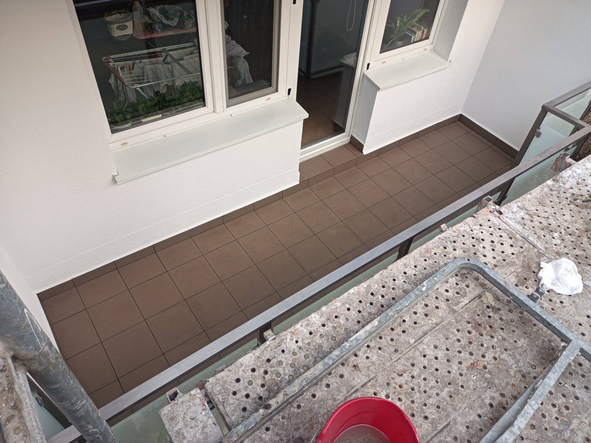 Widok z góry na balkon z brązowymi płytkami, świeżo pomalowanymi na biało ścianami i ramami okiennymi; na pierwszym planie fragment rusztowania z czerwonym wiadrem i białą folią malarską.