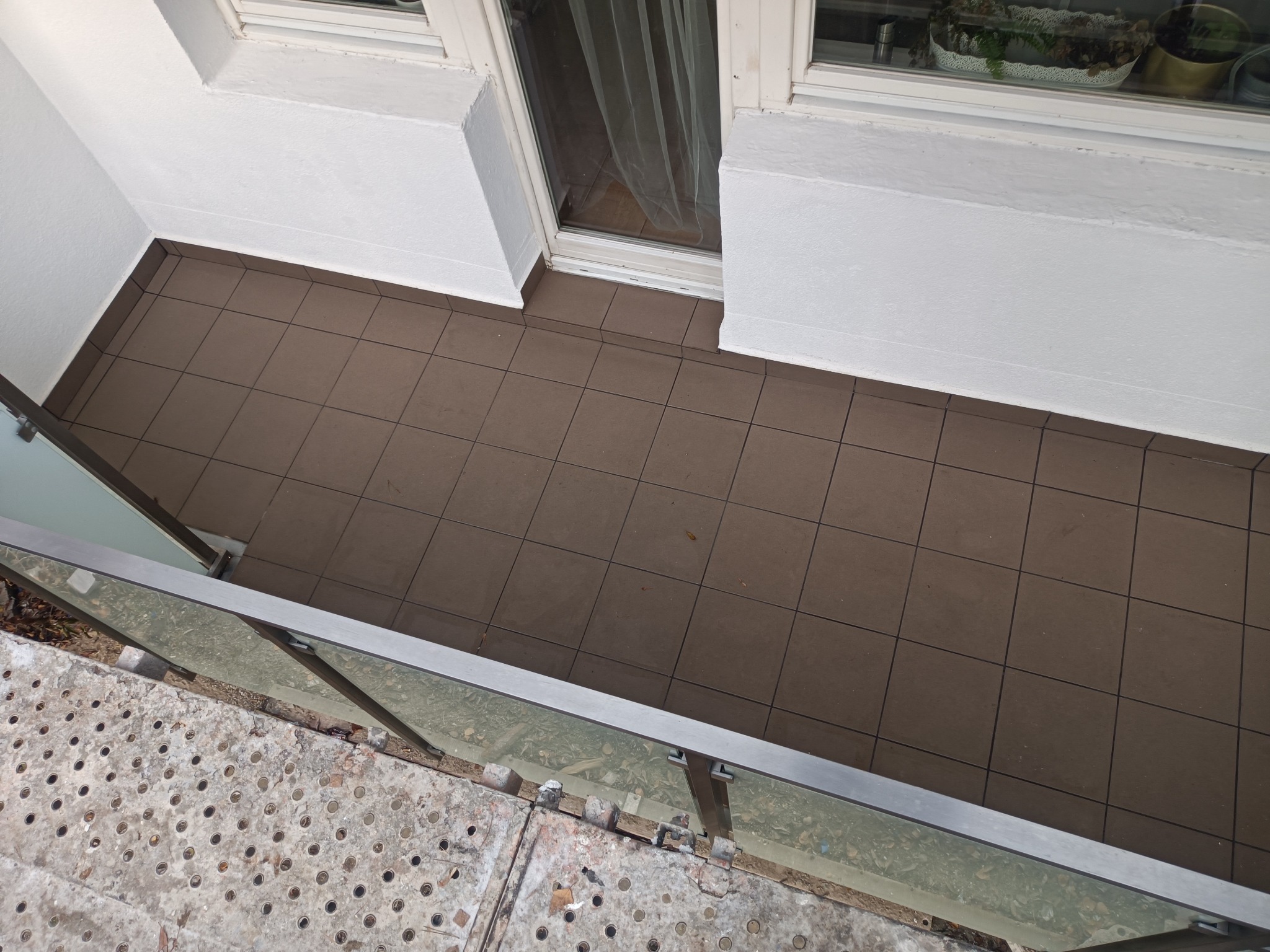 Brązowe płytki na balkonie, białe ściany, szklana balustrada, widok z góry ze schodów lub rusztowania.