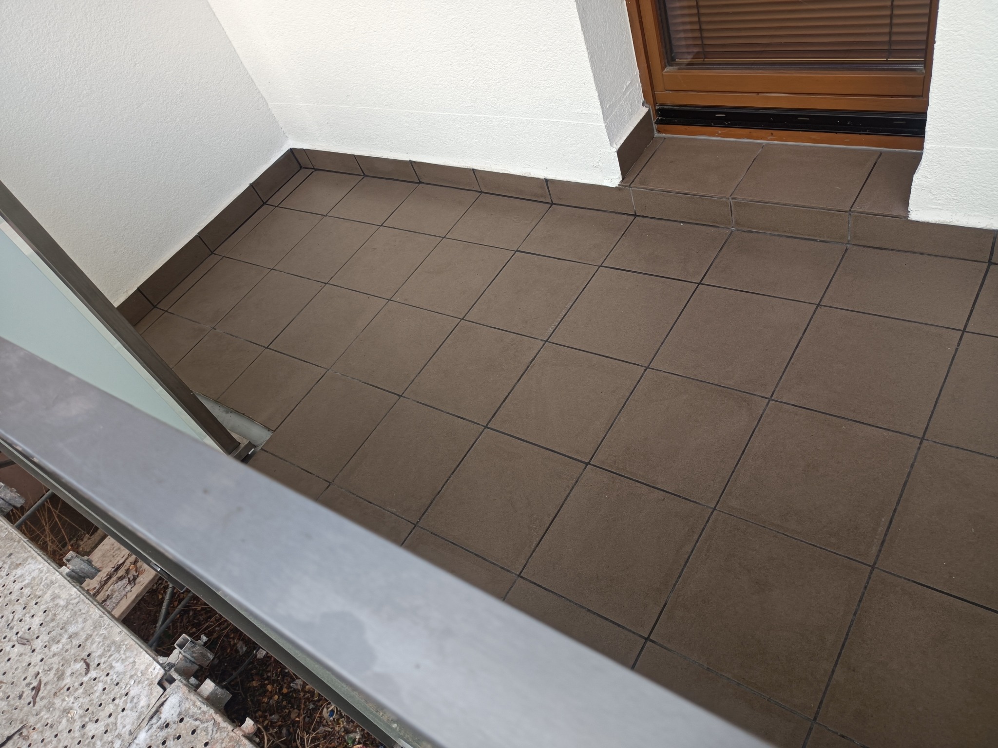 Wykończony balkon z brązowymi płytkami podłogowymi ułożonymi w kwadraty, białe ściany i fragment balustrady w kadrze.