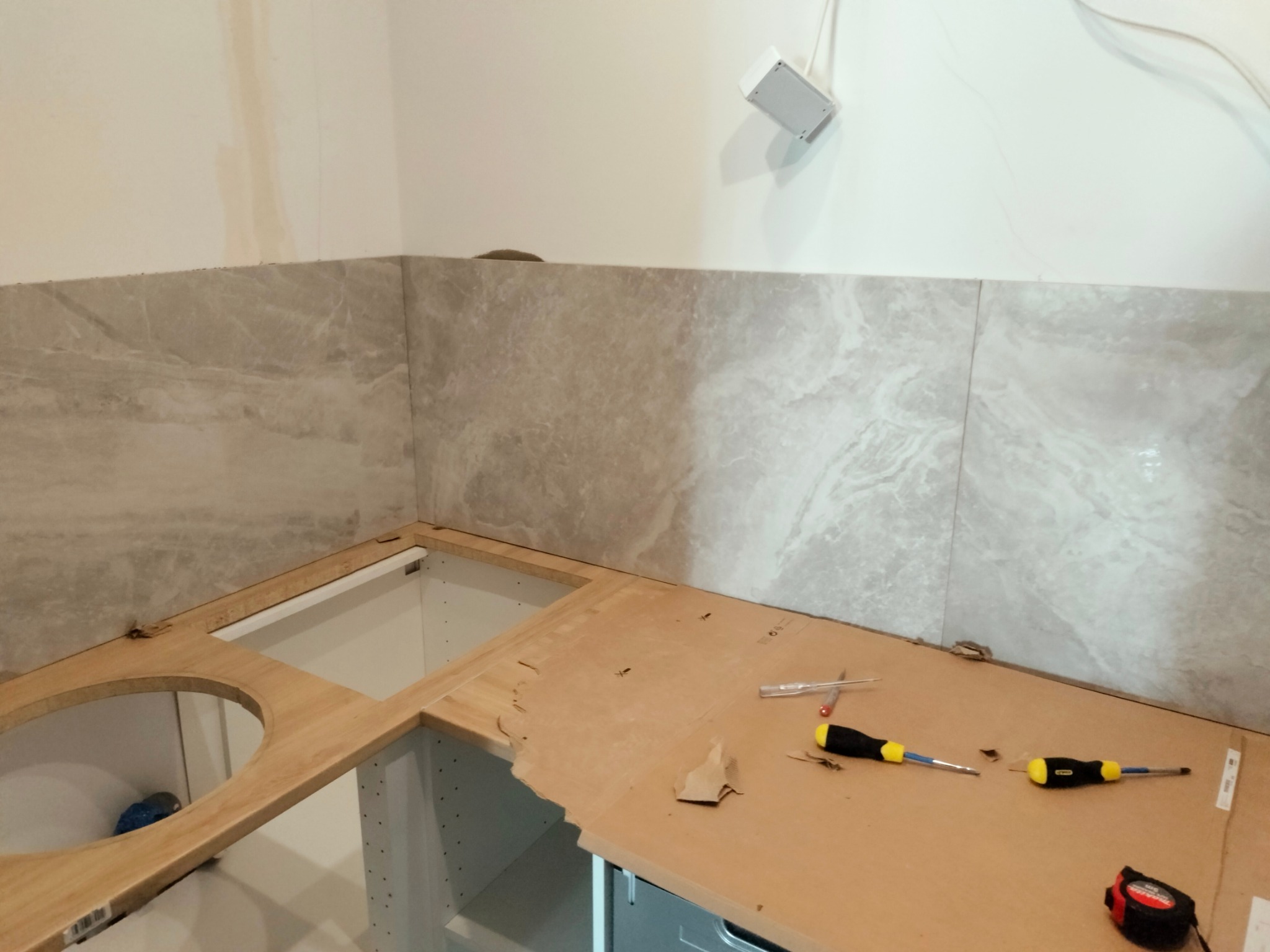 Montaż blatu kuchennego z wyciętym otworem na zlew, częściowo wyłożonego szarymi płytkami imitującymi marmur, z widocznymi narzędziami: dwoma śrubokrętami i miarką.