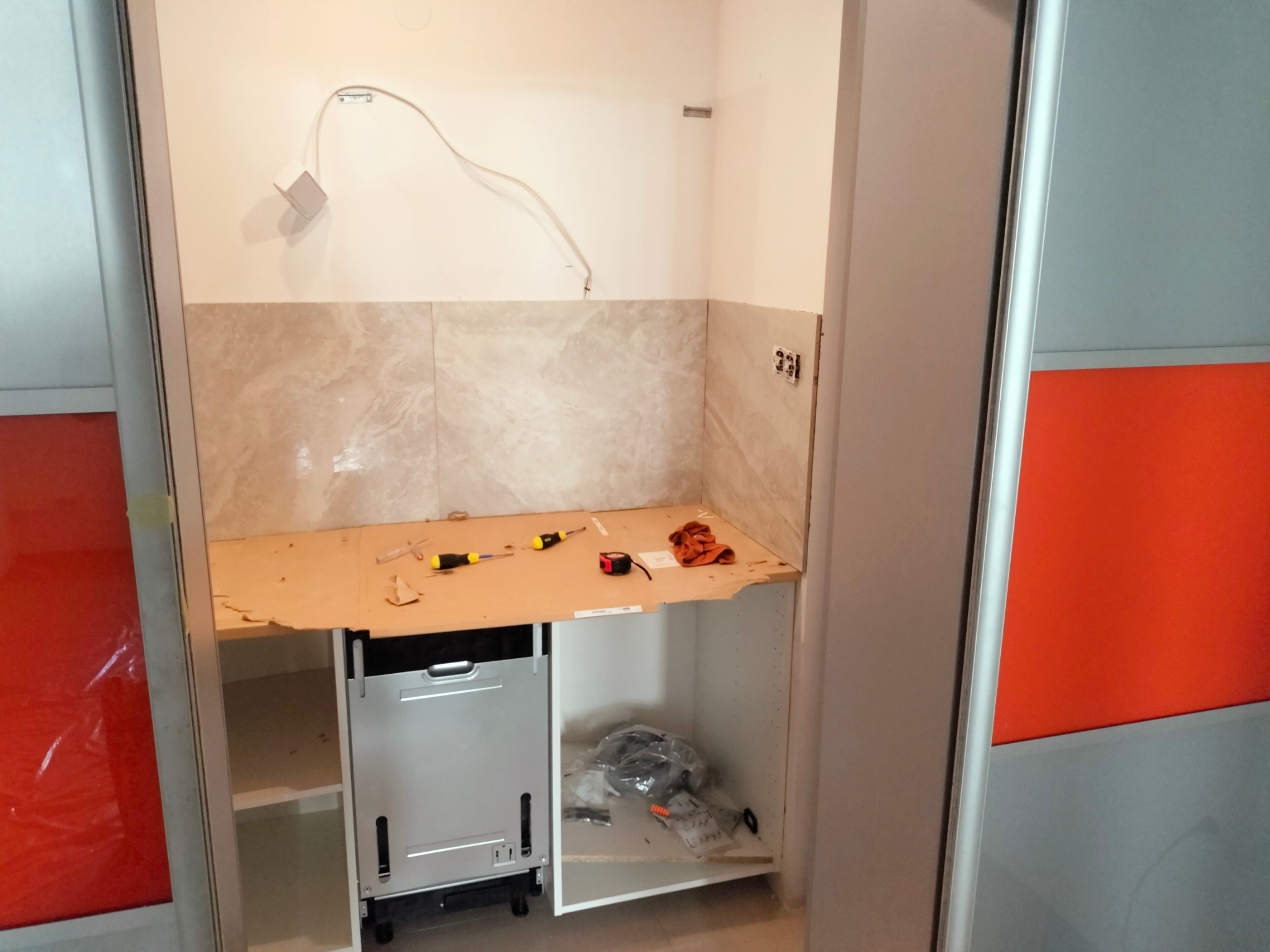 Wnęka w trakcie aranżacji z zamontowanymi płytkami imitującymi marmur, przygotowana pod zabudowę kuchenną ze sprzętem AGD, widoczne narzędzia i kable elektryczne