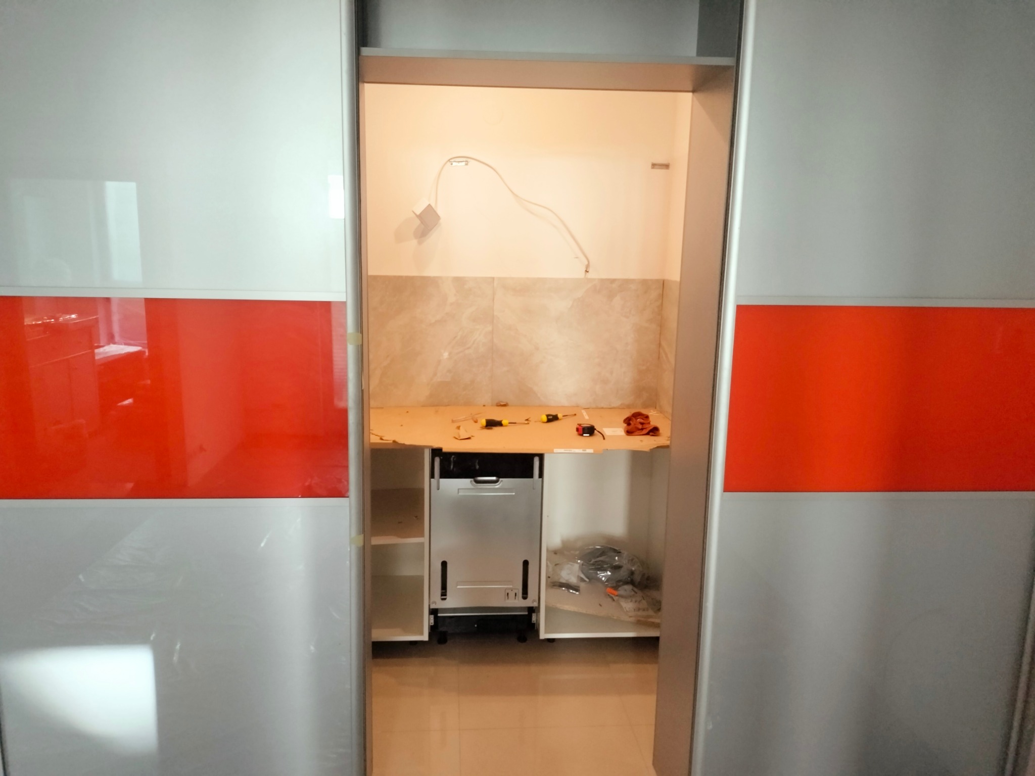 Wnęka w trakcie przygotowania do montażu zmywarki, widoczne narzędzia, instalacja elektryczna i częściowo ułożone płytki na ścianie, przesuwne drzwi z pomarańczowym pasem po bokach.