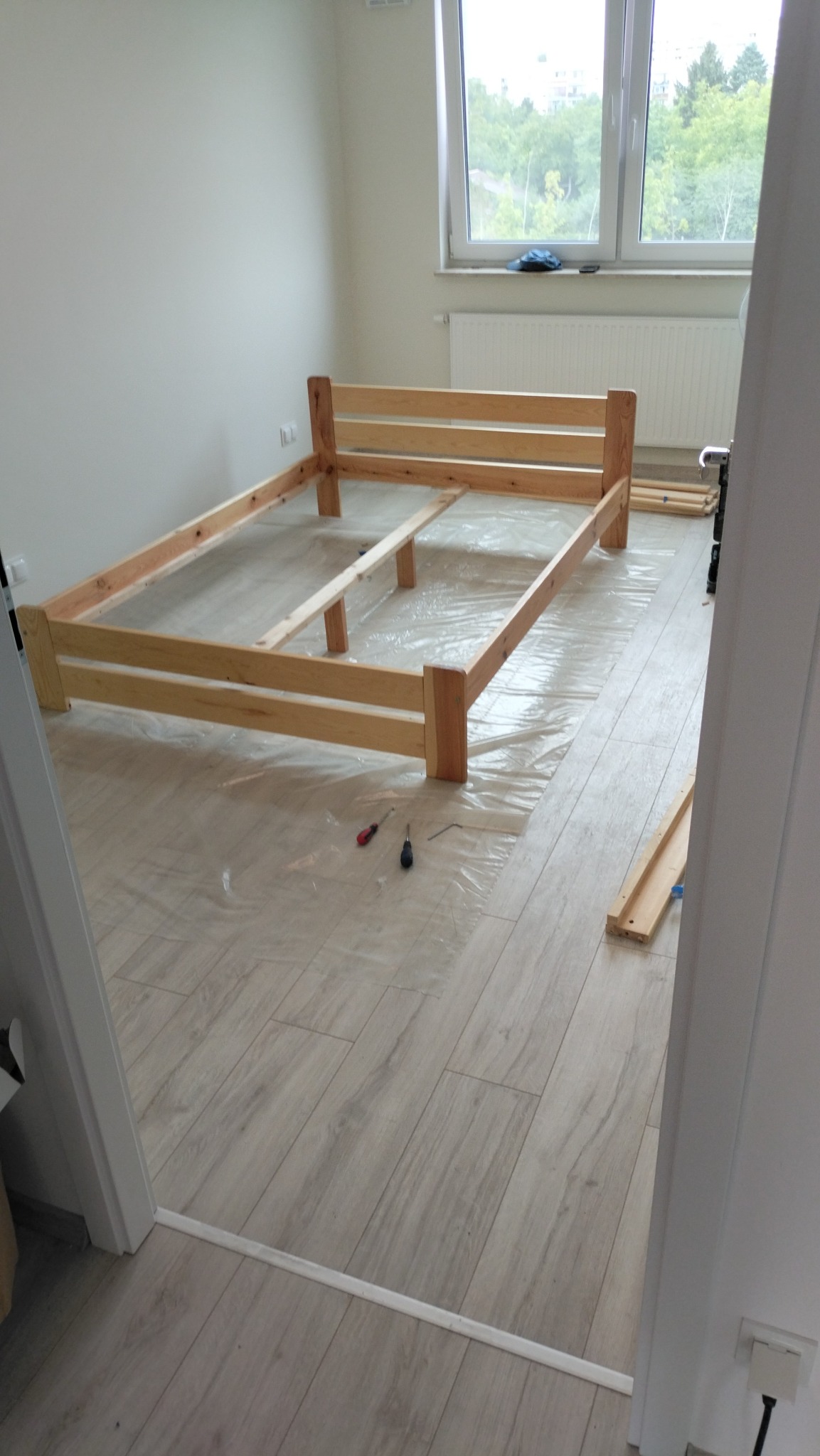 Rama łóżka z jasnego drewna w trakcie montażu, zabezpieczona folią na podłodze z płytek imitujących drewno, widoczne narzędzia i fragmenty ramy.