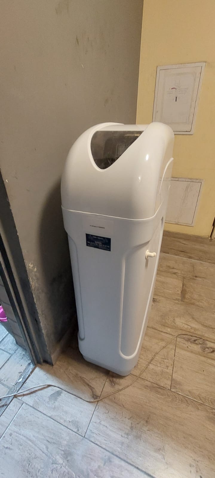 Biały kabinet do zmiękczania wody, ustawiony w rogu pomieszczenia na drewnopodobnych płytkach, z widocznym fragmentem ściany w kolorze beżowym i szarym.