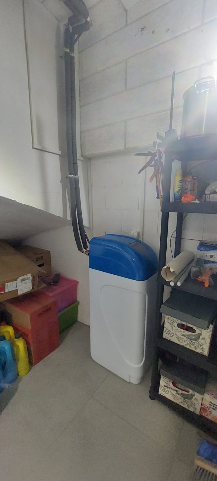 Zainstalowany kompaktowy zmiękczacz wody z niebieską głowicą w pomieszczeniu gospodarczym, widoczne rury doprowadzające wodę i półka z akcesoriami.