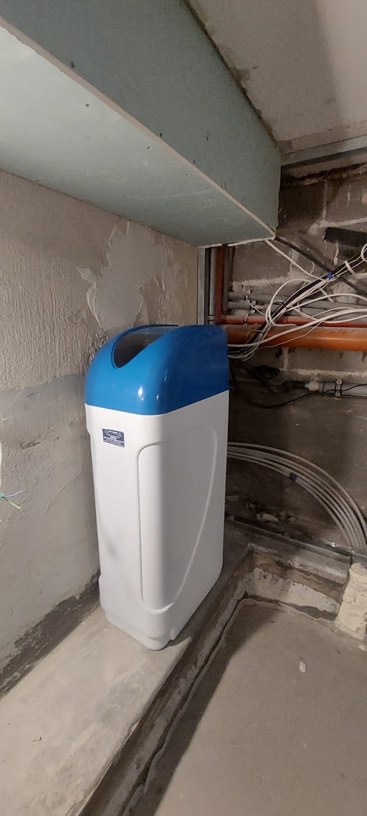 Kompaktowy zmiękczacz wody z niebieską pokrywą, umieszczony w narożniku pomieszczenia w trakcie prac instalacyjnych, widoczne rury i przewody.