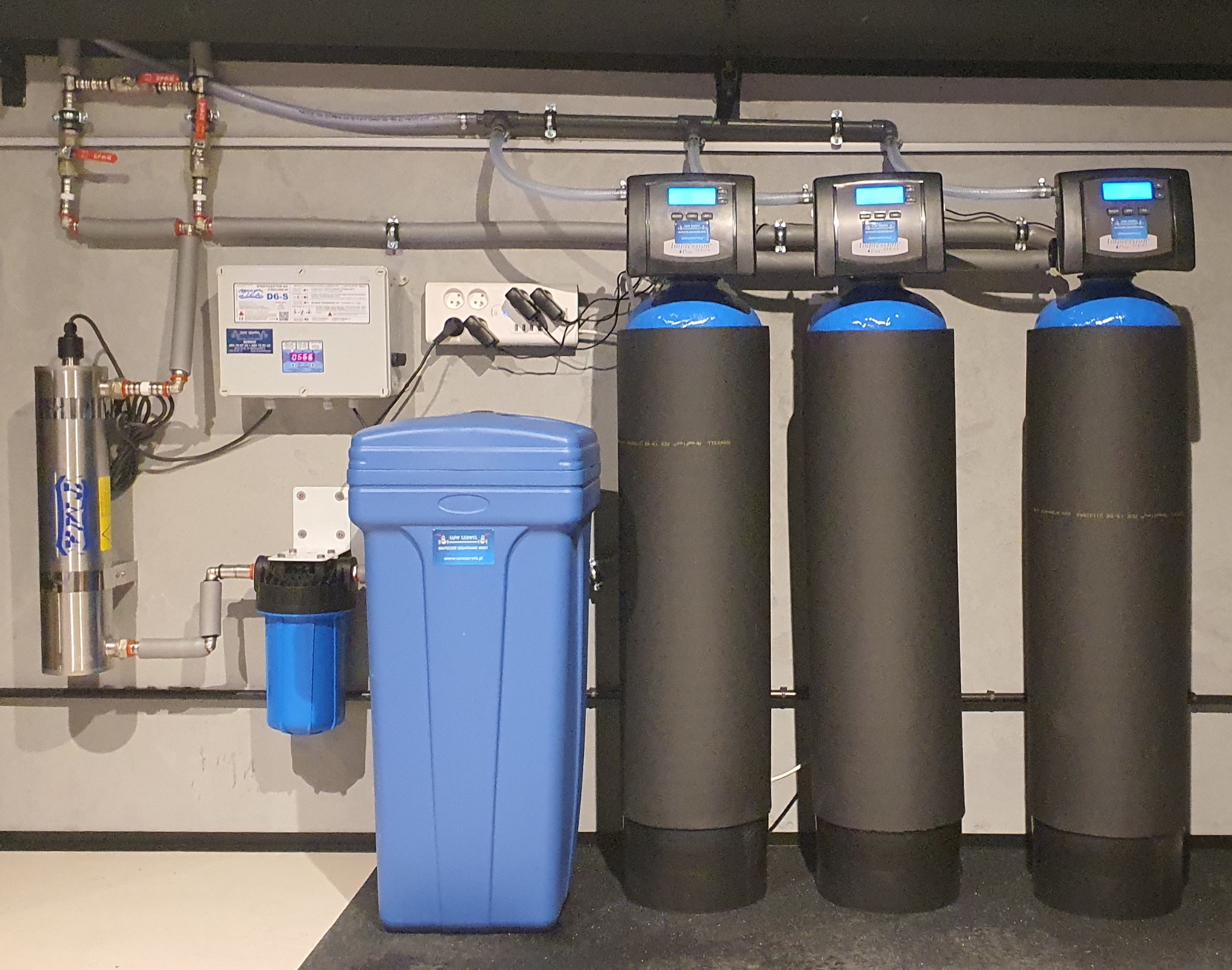 Kompletna instalacja do filtracji wody w domu: lampa UV, filtr narurowy, zmiękczacz wody i trzy kolumny filtracyjne z elektronicznymi sterownikami, zamontowane na szarej ścianie.