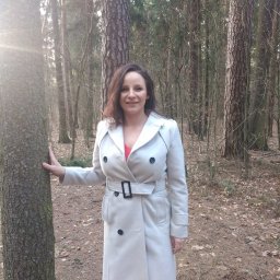 Anna Wilczyńska - Olsztyńskie Centrum Terapii TSR Punkt Zwrotny - Kobieta w jasnym płaszczu opiera dłoń o korę drzewa w lesie, promienie słońca przebijają się przez gałęzie w tle.