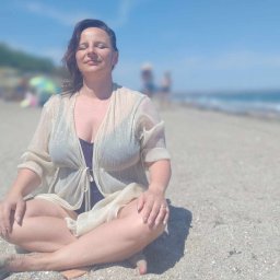 Anna Wilczyńska - Olsztyńskie Centrum Terapii TSR Punkt Zwrotny - Kobieta siedząca w pozycji lotosu na piaszczystej plaży, ubrana w prześwitującą narzutkę i ciemny strój kąpielowy, z zamkniętymi oczami i uśmiechem na twarzy, w tle błękitne morze i niebo.