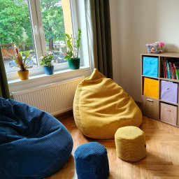 Anna Wilczyńska - Olsztyńskie Centrum Terapii TSR Punkt Zwrotny - Jasne wnętrze z dwoma dużymi pufami (niebieskim i żółtym), małymi pufami, regałem z kolorowymi pojemnikami i roślinami na parapecie, tworzące komfortową przestrzeń.