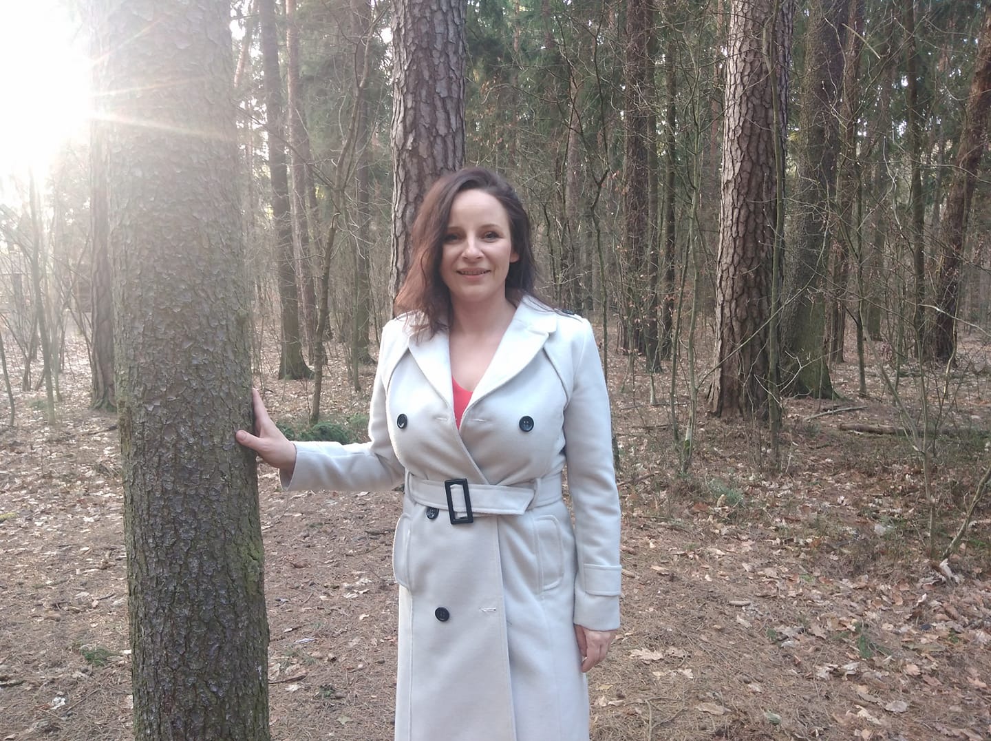 Kobieta w jasnym płaszczu opiera dłoń o korę drzewa w lesie, promienie słońca przebijają się przez gałęzie w tle.