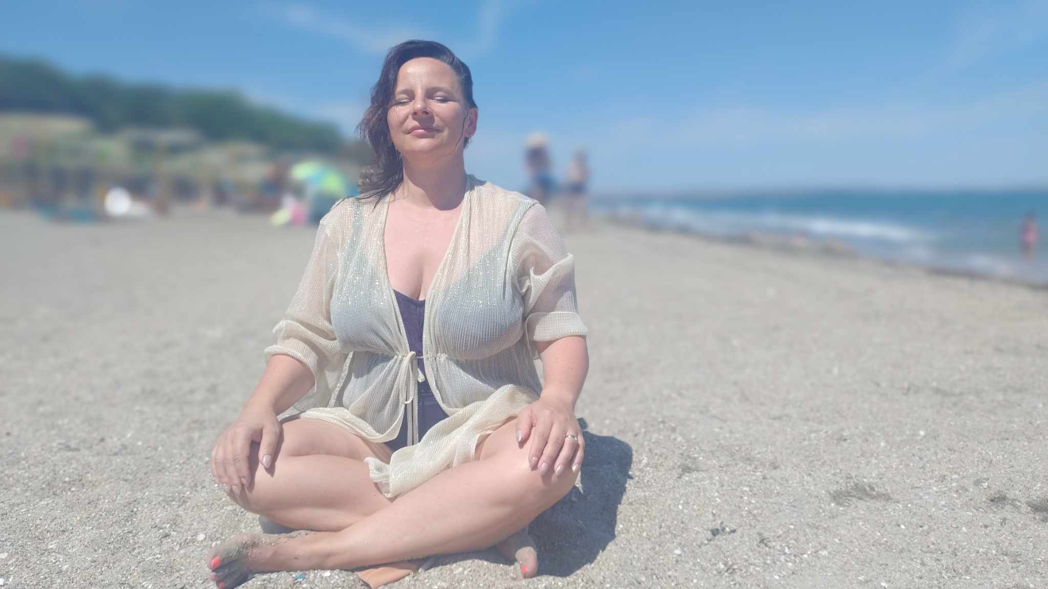 Kobieta siedząca w pozycji lotosu na piaszczystej plaży, ubrana w prześwitującą narzutkę i ciemny strój kąpielowy, z zamkniętymi oczami i uśmiechem na twarzy, w tle błękitne morze i niebo.