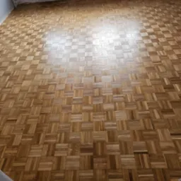 Parkiet ułożony w kwadraty, widok z góry na błyszczącą drewnianą podłogę, fragment grzejnika i buta w rogu kadru.