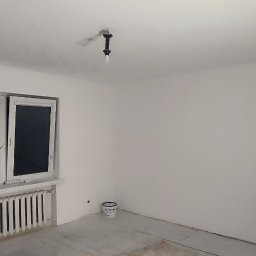 Art-Decor Mariusz Lorenc - Pomalowane na biało wnętrze pokoju z grzejnikiem pod oknem, widoczna puszka z farbą na podłodze i surowa żarówka na suficie.