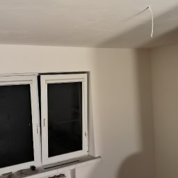 Art-Decor Mariusz Lorenc - Świeżo pomalowane wnętrze pokoju z dwoma oknami. Na suficie widoczne wystające kable elektryczne.