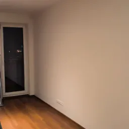 Pusty pokój z brązowymi panelami podłogowymi, świeżo pomalowanymi na biało ścianami i białymi listwami przypodłogowymi, widoczne gniazdka elektryczne i para czarnych butów w rogu.