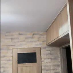 Fragment wnętrza po odświeżeniu: drewnopodobne panele ścienne w odcieniach beżu i szarości, drewniane drzwi z czarną szybą, oraz wisząca szafka w kolorze jasnego drewna, widoczne z perspektywy dolnej.