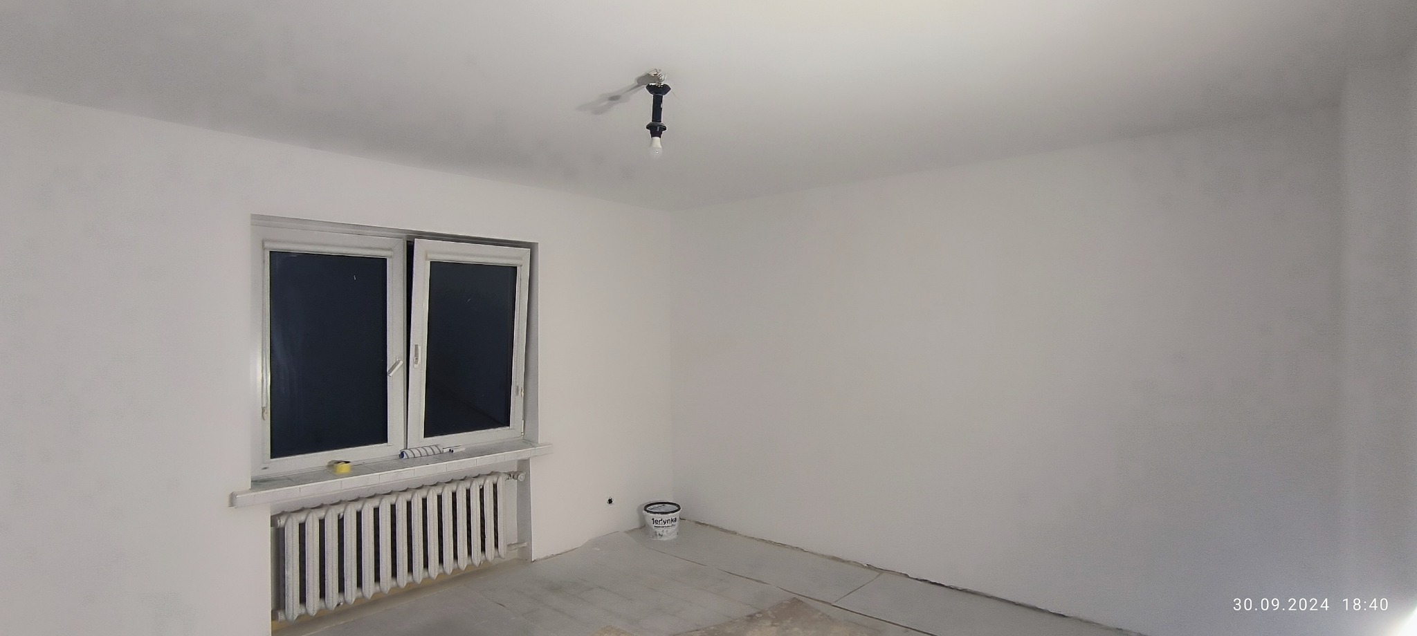 Pomalowane na biało wnętrze pokoju z grzejnikiem pod oknem, widoczna puszka z farbą na podłodze i surowa żarówka na suficie.