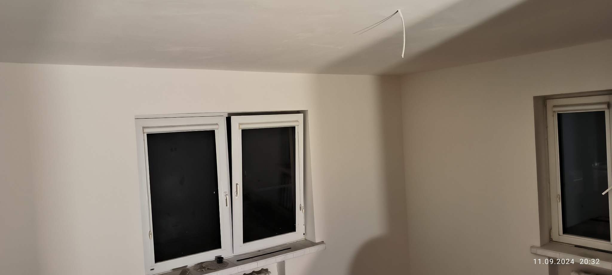 Świeżo pomalowane wnętrze pokoju z dwoma oknami. Na suficie widoczne wystające kable elektryczne.