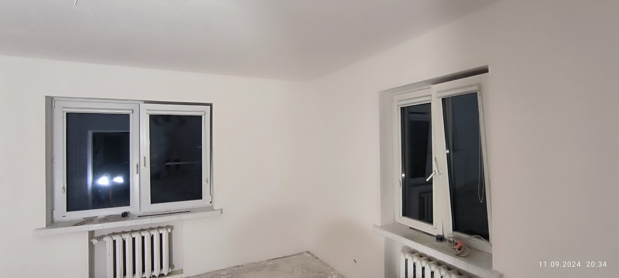 Pokój po malowaniu na biało, z dwoma oknami PCV, grzejnikiem żeliwnym i widocznymi śladami prac na podłodze.