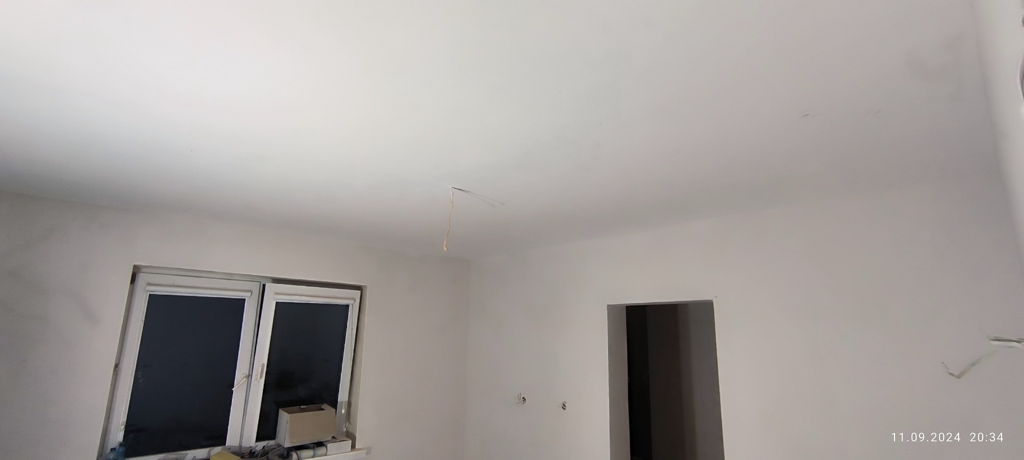 Wnętrze w trakcie prac wykończeniowych: gładkie, białe ściany i sufit, widoczne okno z roletami oraz otwór drzwiowy, z sufitu zwisa kabel.