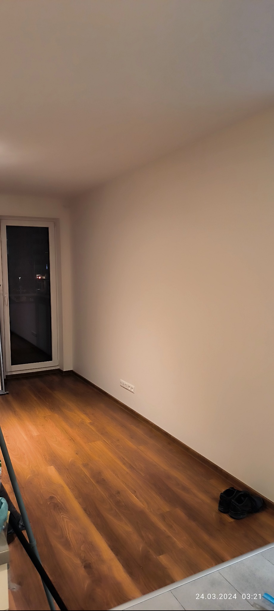 Pusty pokój z brązowymi panelami podłogowymi, świeżo pomalowanymi na biało ścianami i białymi listwami przypodłogowymi, widoczne gniazdka elektryczne i para czarnych butów w rogu.