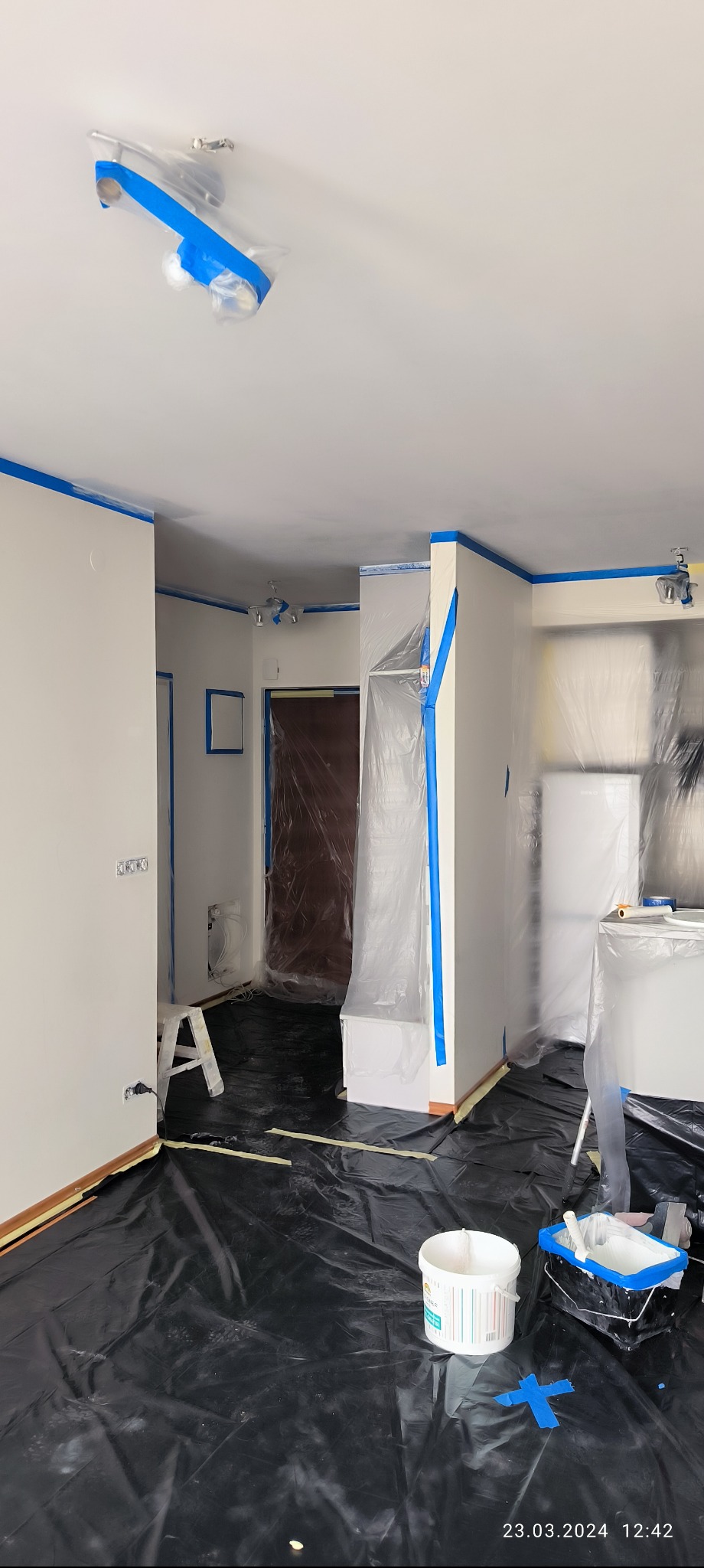 Przygotowania do malowania mieszkania: pomieszczenie zabezpieczone folią i taśmą malarską, widoczne wiaderko z farbą i drabina.