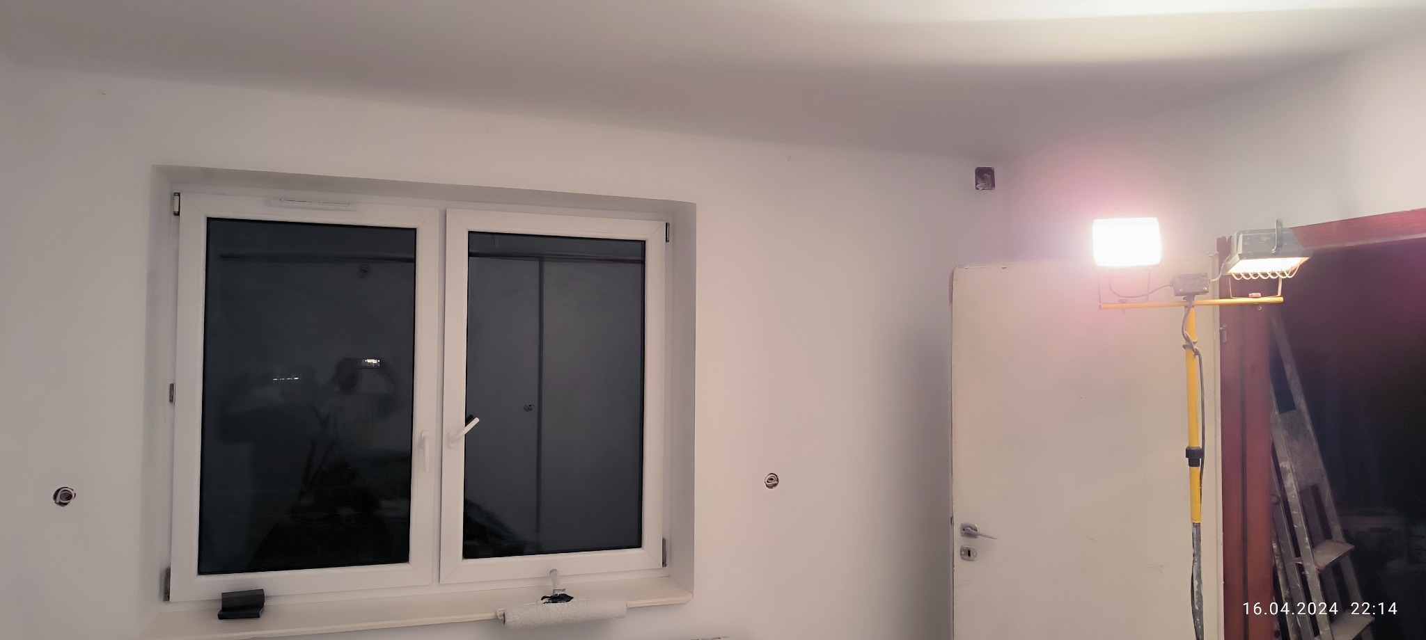 Białe ściany w trakcie prac, widoczne przygotowane otwory pod gniazdka elektryczne, okno z roletami, lampa budowlana na statywie i drabina oparta o ścianę.