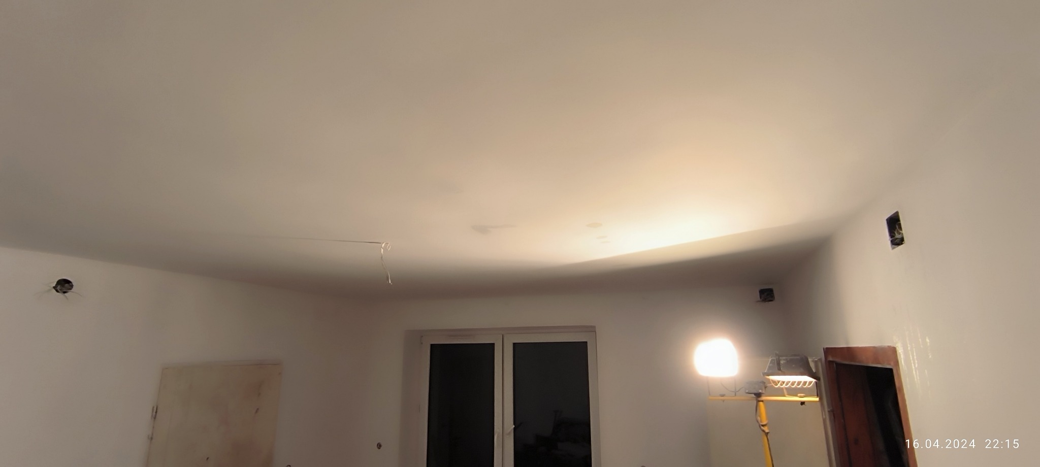 Pokój w trakcie prac wykończeniowych, widoczna lampa robocza, otwory w ścianach i wystające kable elektryczne, częściowo pomalowane ściany i sufit.