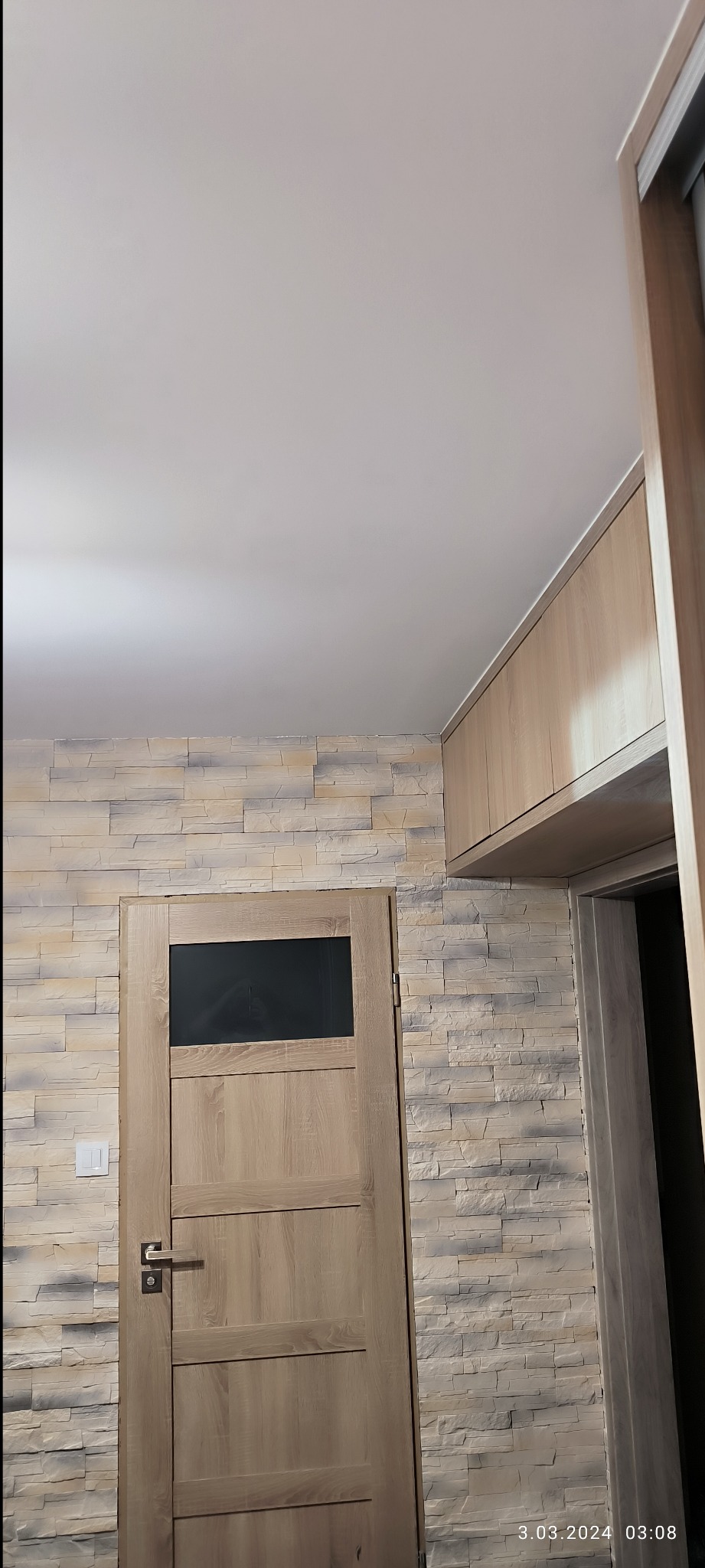 Fragment wnętrza po odświeżeniu: drewnopodobne panele ścienne w odcieniach beżu i szarości, drewniane drzwi z czarną szybą, oraz wisząca szafka w kolorze jasnego drewna, widoczne z perspektywy dolnej.