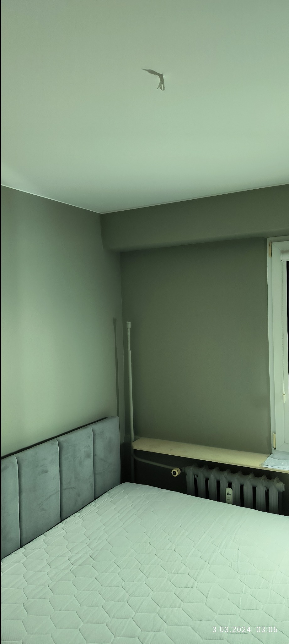 Pokój z łóżkiem, szarą ścianą i grzejnikiem, widoczne ślady po taśmie na suficie.