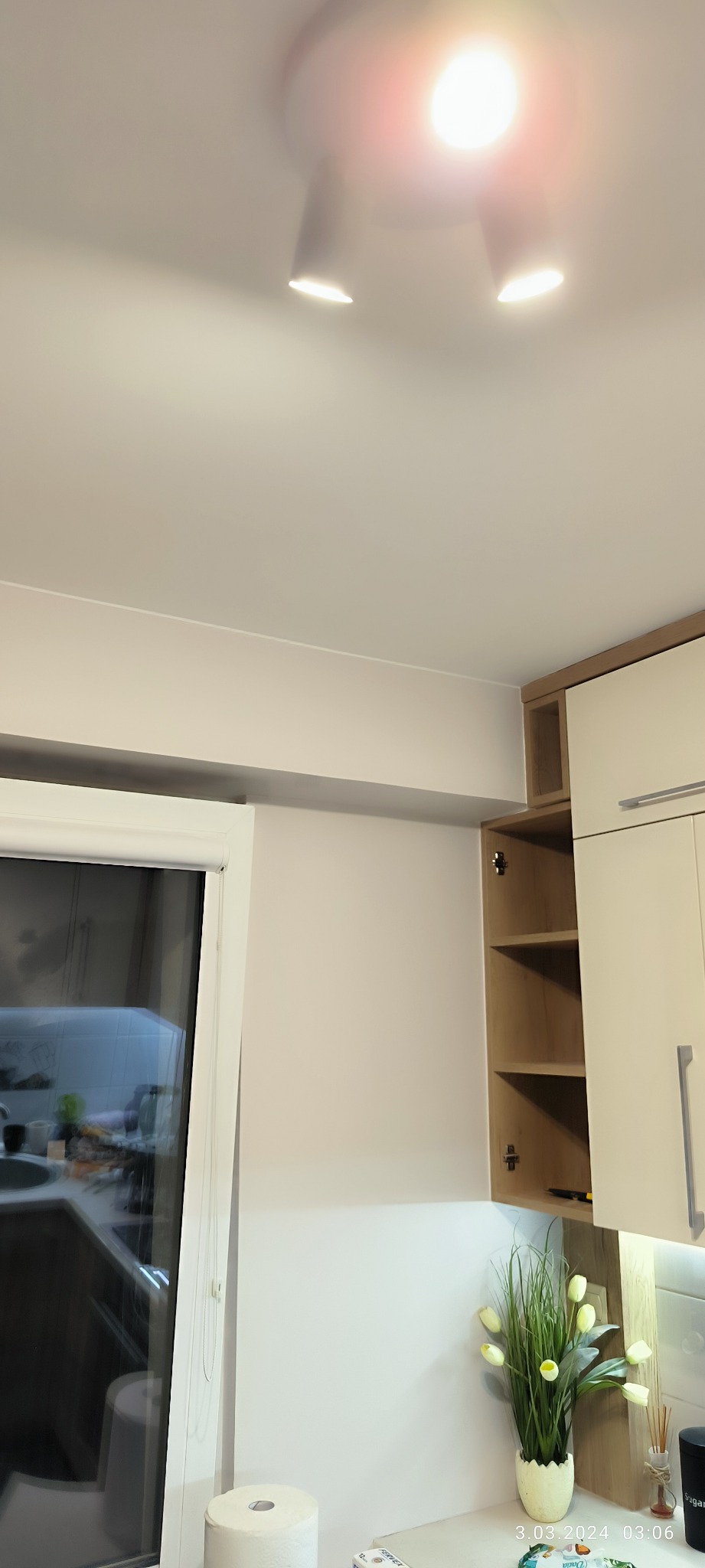 Nowoczesna kuchnia z białymi szafkami, otwartą półką i sztucznymi tulipanami w doniczce, oświetlona trzema punktowymi lampami sufitowymi.