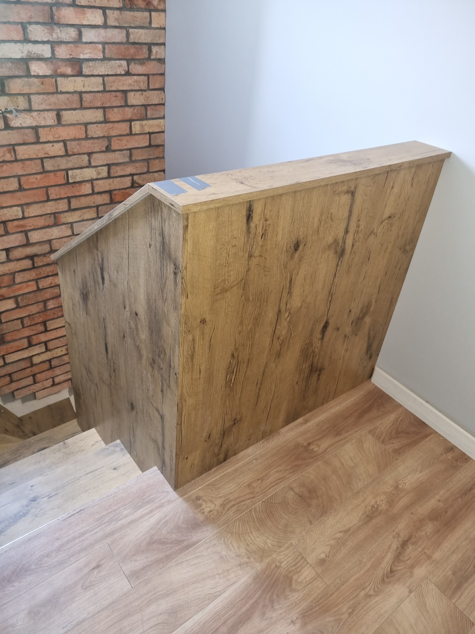 Fragment schodów z drewnianą balustradą o fakturze dębu, widok z góry na stopnie z jasnego drewna, w tle ściana z cegły i biała ściana.