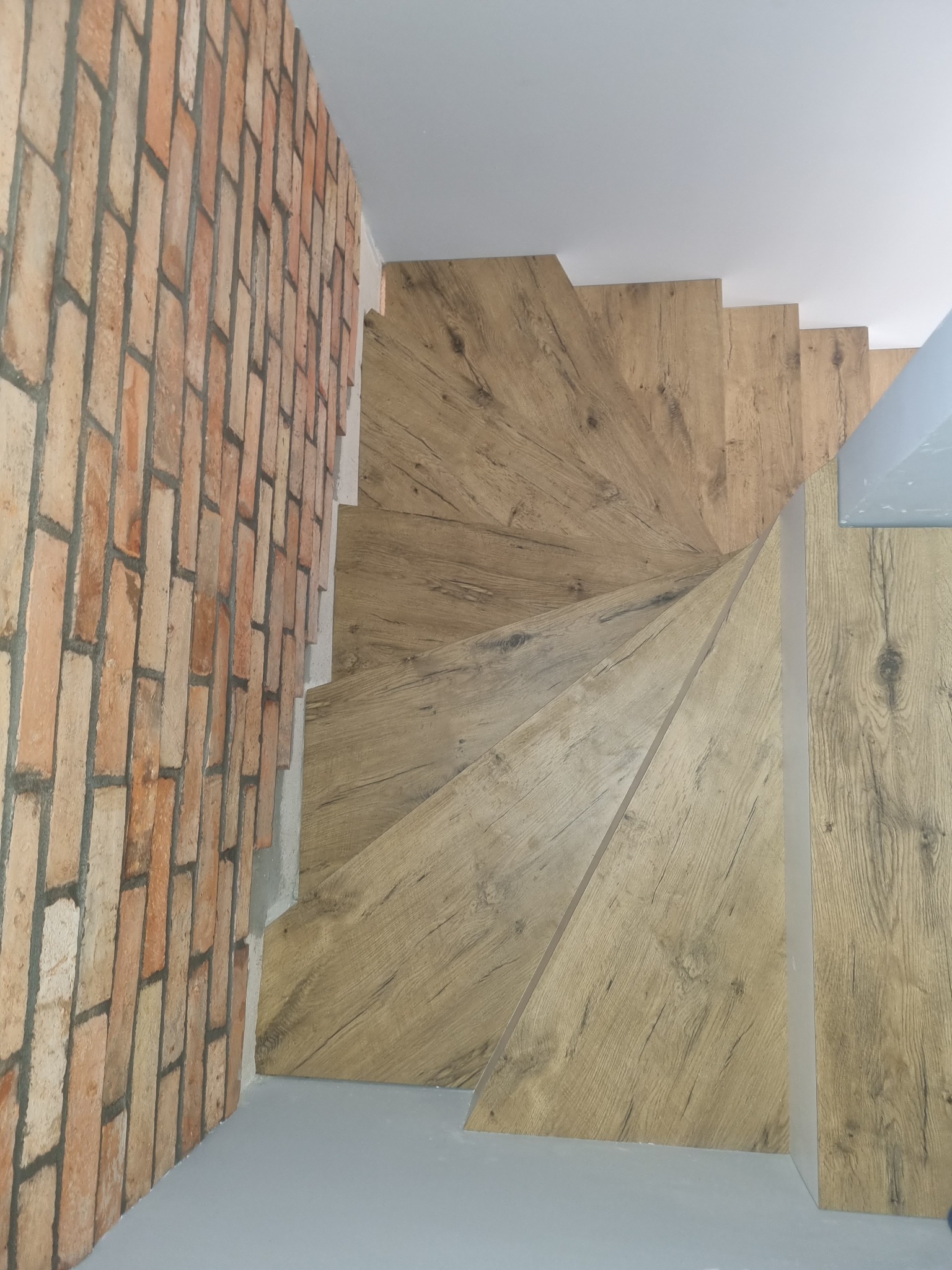 Schody spiralne z drewnopodobnymi panelami, widok z góry, kontrast z ceglaną ścianą. Stopnie o jasnym odcieniu drewna układają się w spiralę, przylegając do ściany z czerwonej cegły.