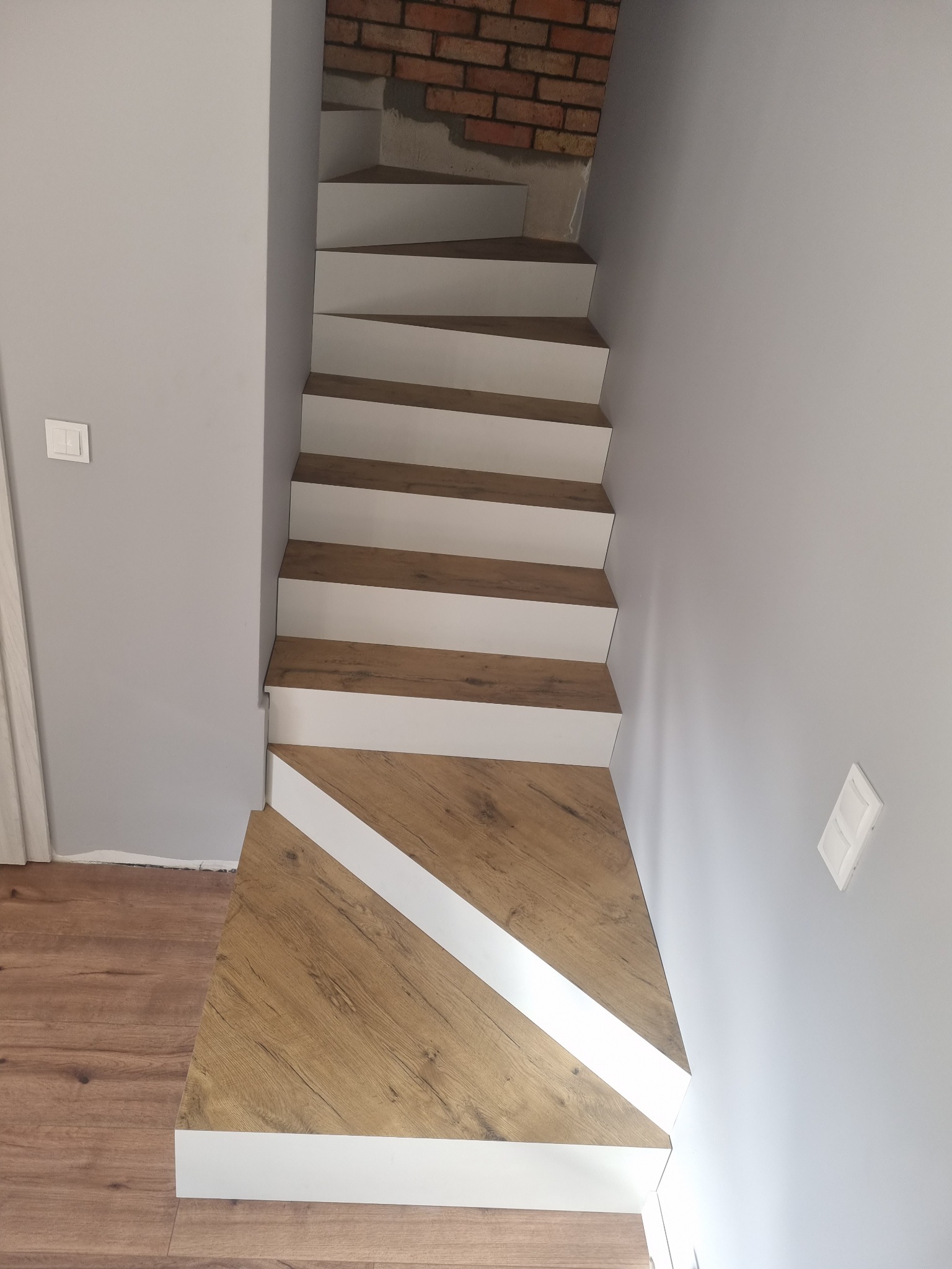 Nowoczesne schody drewniane z białymi podstopnicami, widok z dołu, z fragmentem ściany z cegły w tle.