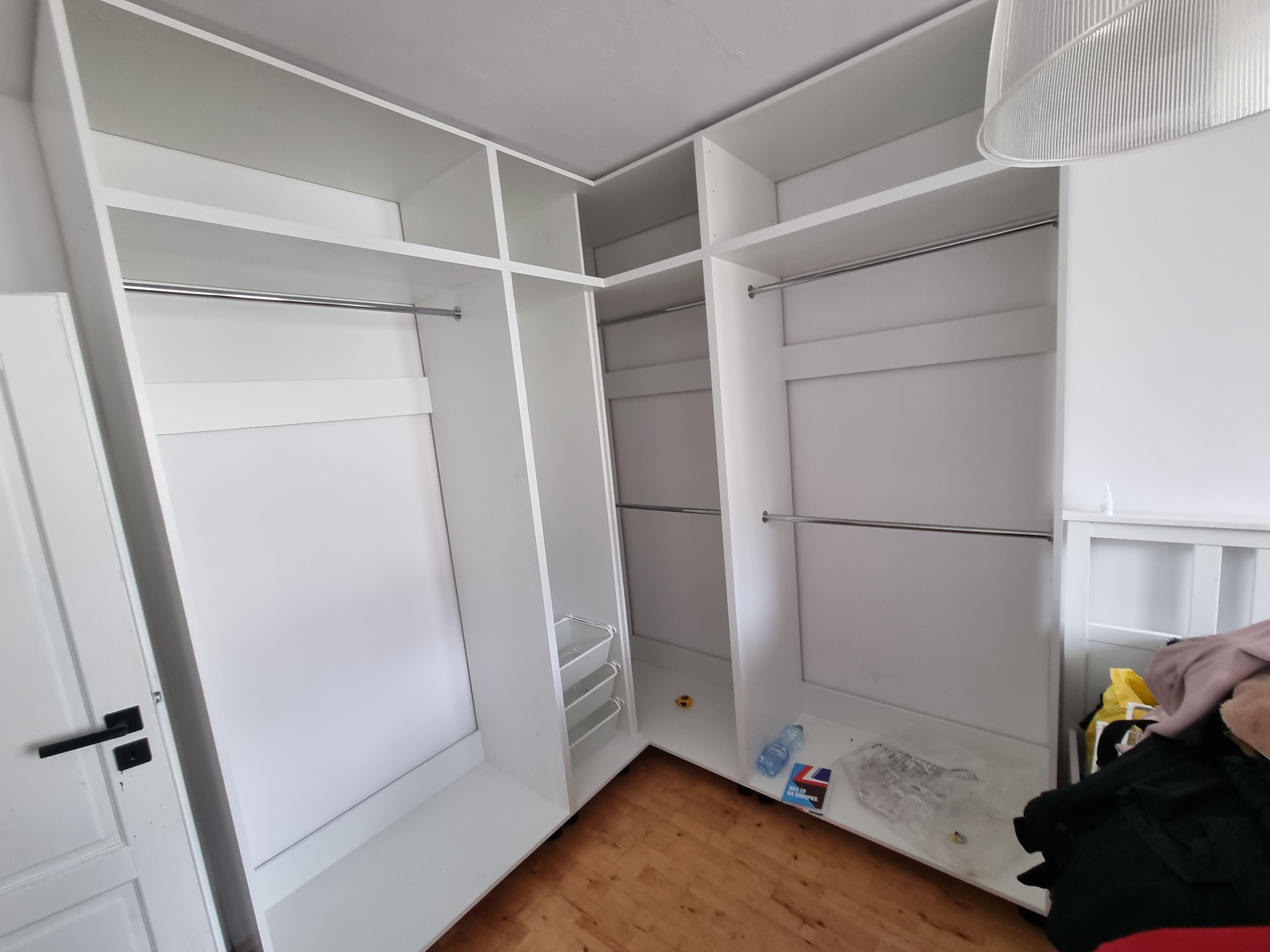 Wnętrze białej, narożnej szafy wnękowej z drążkami na ubrania i wysuwanymi koszami, widoczne niedoskonałości wykończenia i drobne przedmioty na podłodze.
