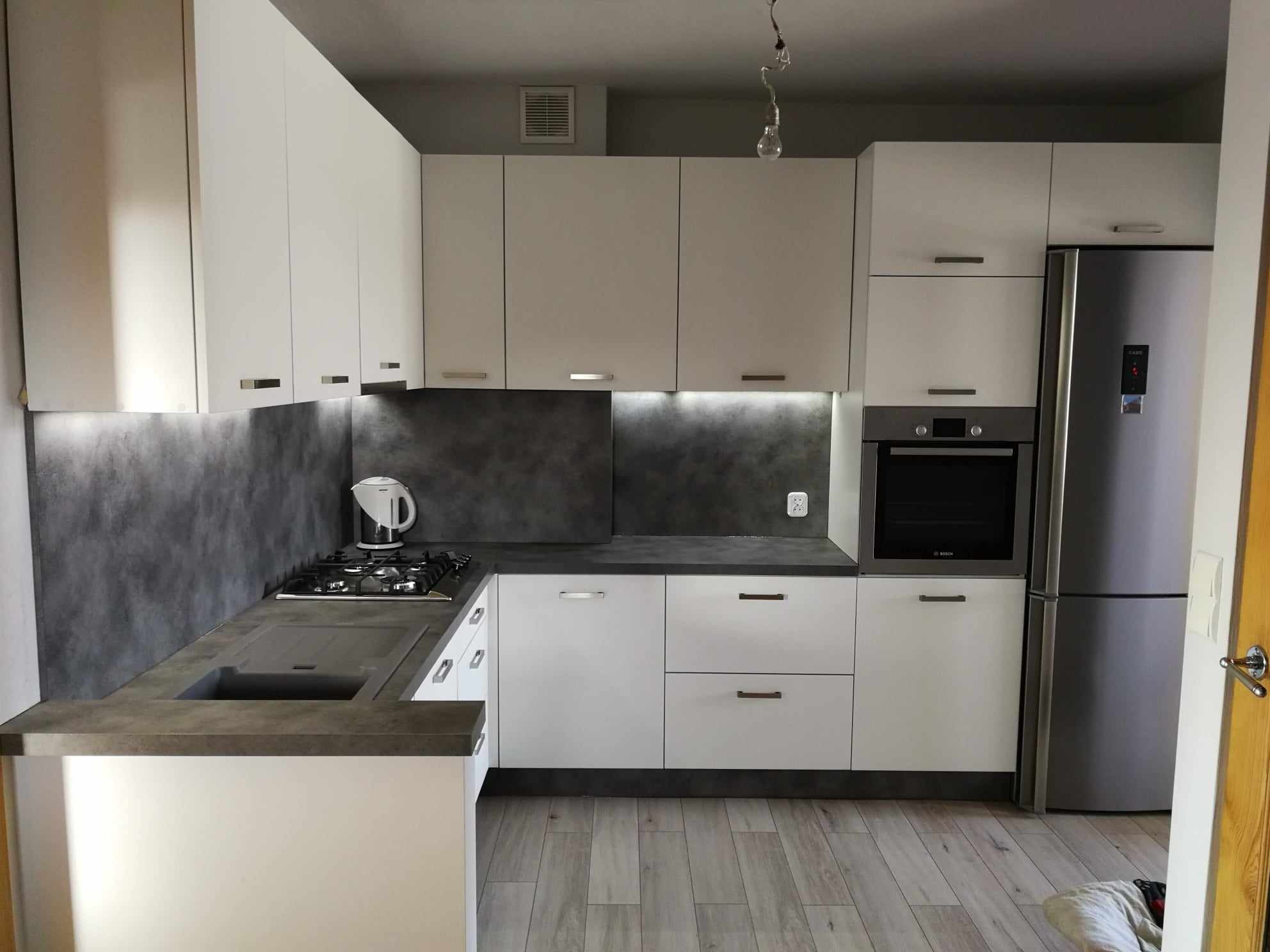 Nowoczesna kuchnia z białymi szafkami, szarym blatem i płytkami imitującymi beton, oświetleniem LED pod szafkami wiszącymi oraz srebrną lodówką, widziana z perspektywy pod lekkim kątem.