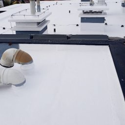 IZO PERFECT Mateusz Gardoliński - Biały, płaski dach pokryty nową powłoką hydroizolacyjną, z widocznymi dwoma rurami wentylacyjnymi i elementami wentylacji, w tle budynki.