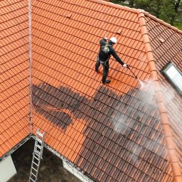 IZO PERFECT Mateusz Gardoliński - Czerwona dachówka czyszczona myjką ciśnieniową przez pracownika w uprzęży, widok z góry. Część dachu umyta, część brudna. Obok drabina i okna dachowe.