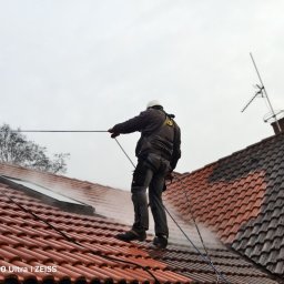 IZO PERFECT Mateusz Gardoliński - Pracownik w uprzęży czyści dachówkę pod ciśnieniem, widoczna różnica w czystości połaci. Antena na kominie w tle, pochmurne niebo.