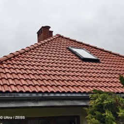 IZO PERFECT Mateusz Gardoliński - Czerwona dachówka ceramiczna z oknem dachowym i kominem z cegły na tle pochmurnego nieba. Widoczne rynny i fragment zieleni.