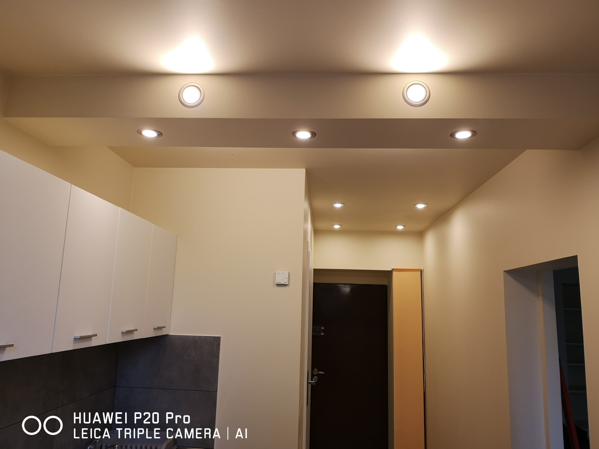 Kuchnia z białymi szafkami, szarym blatem i oświetleniem sufitowym w postaci kilku punktowych lamp.