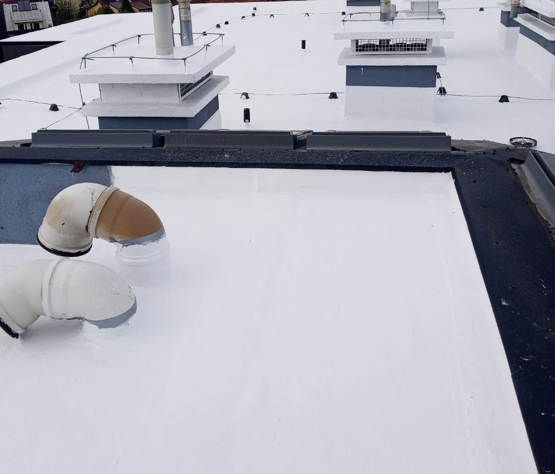 Biały, płaski dach pokryty nową powłoką hydroizolacyjną, z widocznymi dwoma rurami wentylacyjnymi i elementami wentylacji, w tle budynki.