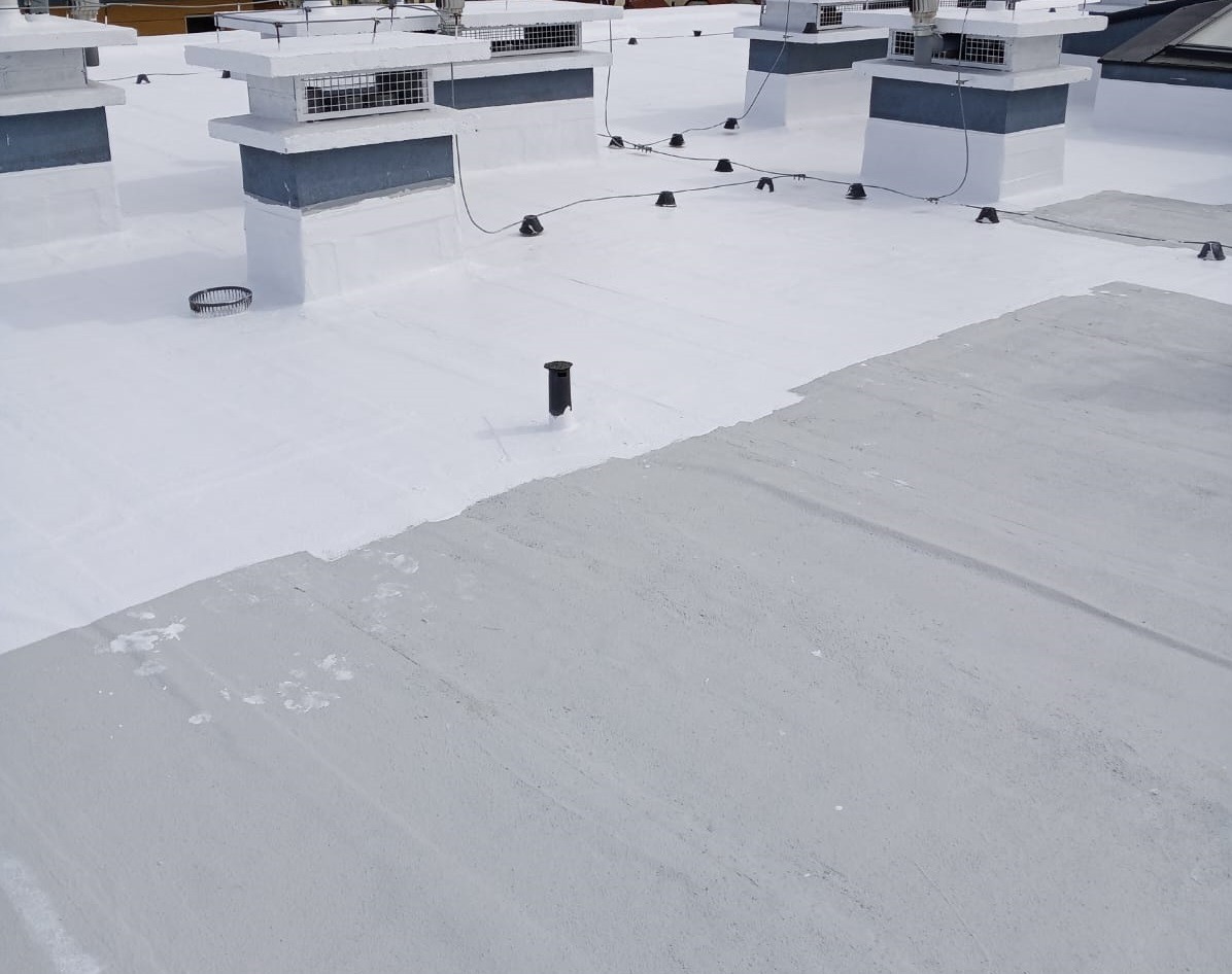 Płaski dach pokryty świeżą, białą powłoką hydroizolacyjną, widoczne kominy wentylacyjne z kratkami i elementy instalacji elektrycznej na tle częściowo pomalowanej powierzchni.